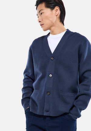 Jeune homme portant un cardigan bleu marine boutonné sur une chemise blanche, les mains dans les poches, regardant sur le côté sur un fond uni.