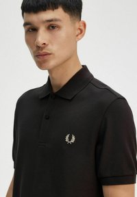 Polo negro hecho de tela texturada, con un collar abotonado y un pequeño logo de laurel bordado en blanco en el lado izquierdo del pecho.