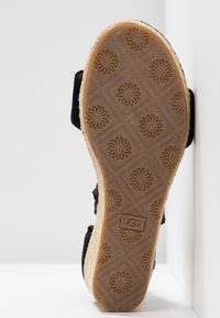 Semelle d'une sandale avec des motifs floraux texturés et le logo "UGG", avec un bord tressé et des lanières noires partiellement visibles.