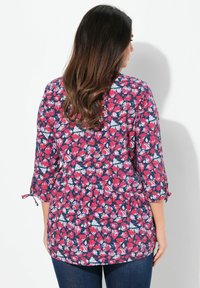 Bloemen tuniektop met marineblauwe basis, met roze en witte bladerenpatronen. Driekwart mouwen met strikdetails, relaxte pasvorm. Spijkerbroek gedragen.