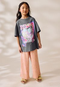 Young girl stands wearing gray oversized disco-themed t-shirt, pink striped wide-leg pants, and beige sandals on neutral background.