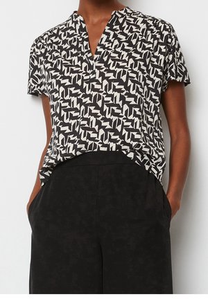 Femme portant un chemisier à manches courtes à motifs noir et blanc, rentré dans un pantalon noir ample taille haute avec les mains dans les poches.