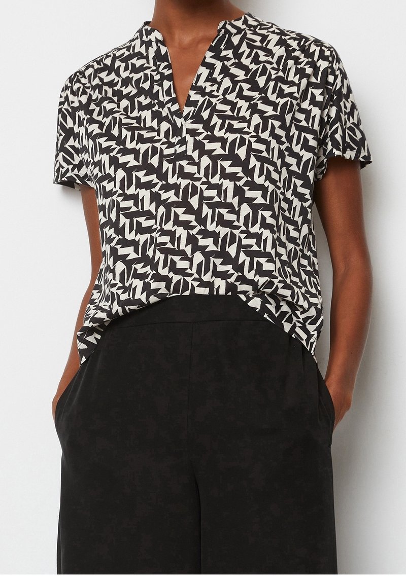 Femme portant un chemisier à manches courtes à motifs noir et blanc, rentré dans un pantalon noir ample taille haute avec les mains dans les poches.