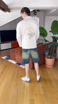 Biela bunda s kvetinovou výšivkou na chrbte, skombinovaná s modrými džínsovými šortkami. Model má na sebe strieborné tenisky. Interiér s rastlinami.