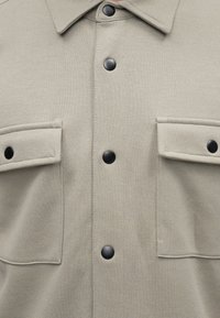 Μπεζ πουκάμισο με υφή και μαύρα κουμπιά με πατήματα στο κέντρο και σε δύο τσέπες με καπάκι στο στήθος, με κωνικό γιακά.