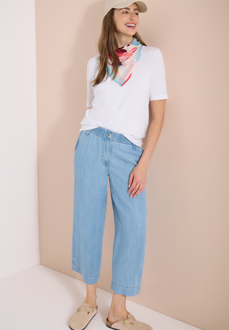 Femme souriante portant une casquette beige, un haut blanc à manches courtes, une écharpe colorée, un pantalon large bleu clair et des chaussures beige sans lacets, debout à l'intérieur.