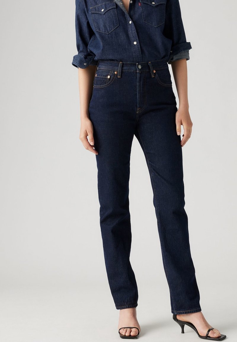 Jeans en denim bleu foncé taille haute avec une jambe droite, cinq poches et des coutures contrastantes, accompagnés d'une chemise en denim foncé et de talons noirs.