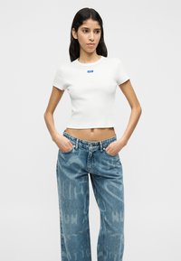 Λευκό κοντό t-shirt με μπλε λογότυπο στο στήθος, συνδυασμένο με ανοιχτό μπλε τζιν παντελόνες με πλατύ πόδι και διακριτικό μοτίβο.