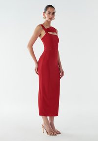 Rød tætsiddende midi-kjole med udskæringer, tykke stropper og en glat tekstur, parret med nude strophæle.