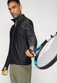 Mustast sportlikust jopest, millel on tõmblukk, tekstuurse paneeliga ja kitsas lõige, kombineeritud oliivroheliste pükstega. Isik hoiab käes tennise reketit ja palli.