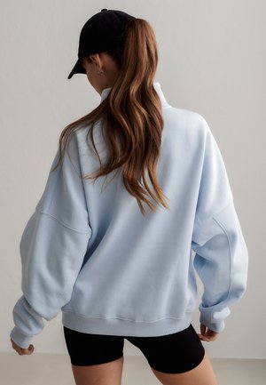 Frau mit langem braunem Haar im Pferdeschwanz, die eine schwarze Mütze, ein hellblaues, übergroßes Sweatshirt und schwarze Shorts trägt und von der Kamera weg schaut.