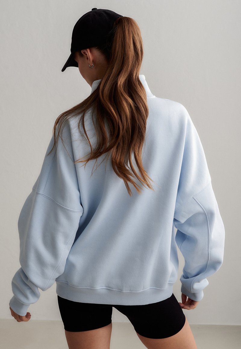 Femme aux longs cheveux bruns attachés en queue de cheval, portant une casquette noire, un sweat-shirt ample bleu clair et un short noir, tournée de dos à la caméra.