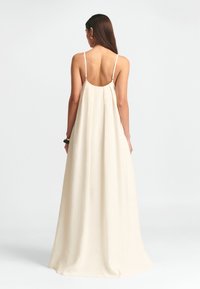 Lang cremefarvet kjole med rygfri design, tynde stropper og en svag silhuet. Stofdet ser blødt ud med en glat tekstur.