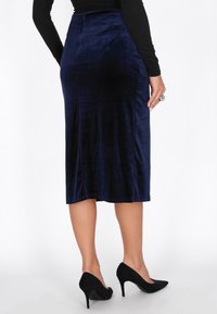 Tengerészkék bársony midi szoknya, testhezálló kialakítással, sima textúrával és hátul slicc részlettel, fekete magas sarkú cipővel párosítva.