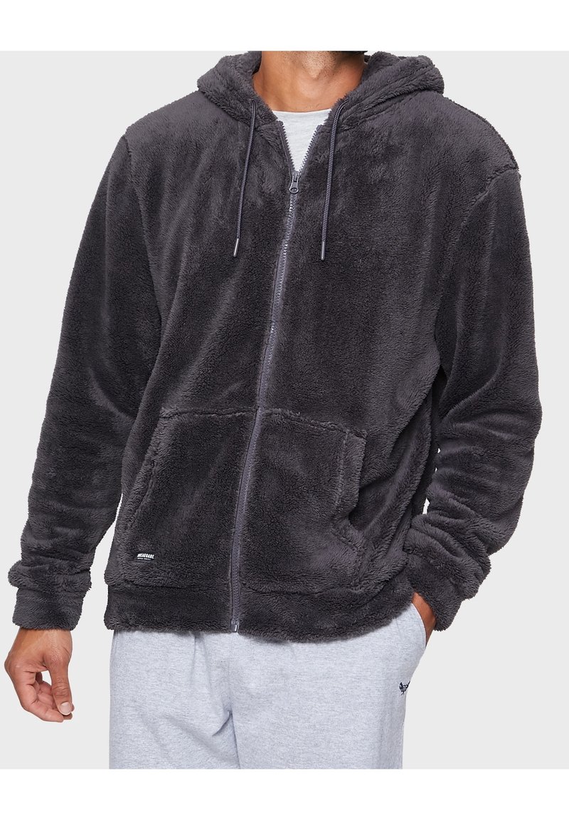 Homme portant un sweat à capuche zippé en peluche gris foncé avec poche avant et pantalon gris, debout avec une main dans la poche sur fond neutre.