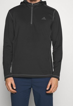 Musta pitkähihainen urheilupaita, jossa on neljännesvetoketju ja Adidaksen logo rinnassa, yhdistetty laivastonsinisiin housuihin.