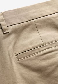 Beige Baumwollhosen mit einer glatten Textur, ausgestattet mit einer Paspeltasche und auffälligen Nähten entlang des Hosenbunds und der Taschenkanten.
