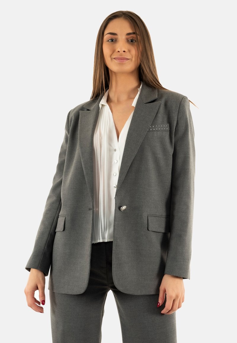 Blazer grigio su misura con revers a punta, chiusura con un bottone e due tasche frontali. Indossato sopra una blusa bianca plissettata. Texture liscia.