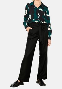 Long black trousers with a tied waist, paired with a green and black patterned blouse featuring a collar and buttons. Smooth fabric texture.