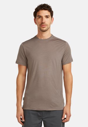 Hombre de pie, mirando al frente, vistiendo una camiseta de manga corta lisa de color marrón claro y pantalones gris oscuro contra un fondo blanco.