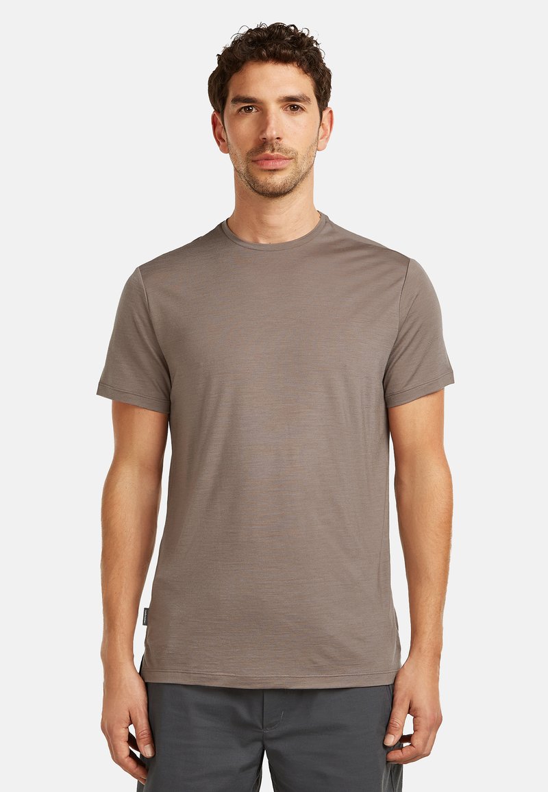 Man staat met het gezicht naar voren, draagt een effen lichtbruin T-shirt met korte mouwen en donkergrijze broek tegen een witte achtergrond.