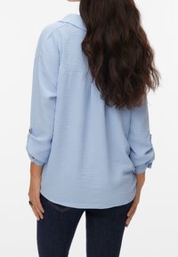 Femme aux longs cheveux ondulés et foncés, portant une chemise texturée bleu clair à manches longues et un jean bleu foncé, vue de dos.