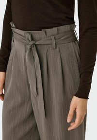 Grijze geplooide broek met een strikband, gemaakt van een textuurstof. De drager heeft een bruine shirt met lange mouwen zichtbaar.