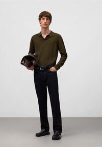 Olivgrünes Polohemd, schwarze Hose und schwarze Schuhe. Hält eine dunkelbraune Tasche. Einfaches Design, strukturierter Stoff und figurbetonte Silhouette.