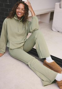 Svetlozelený oversized pulóver s polovičným zipsom a prednou vreckom, skombinovaný s ladnými priliehavými nohavicami s bočnými švami. Hnedé nazúvacie topánky.