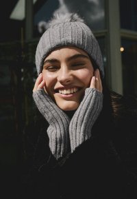 Grijze gebreide muts met een pompon bovenop en bijpassende vingerloze wanten. Het materiaal heeft een geribbelde textuur met subtiele glinsteraccenten.