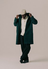 Mørkegrønn rutete frakk med dobbeltradet design, båret over en hvit teksturert genser og svarte bukser, kombinert med svarte sko og en beige caps.