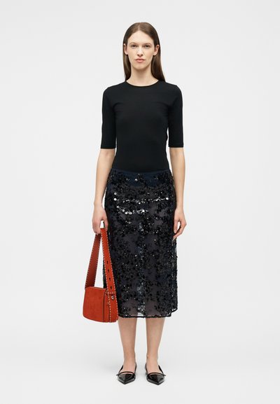 Γυναίκα που φοράει μαύρο τοπ με μανίκια μέχρι τον αγκώνα και μαύρη φούστα με παγιέτες μέχρι τα γόνατα, κρατώντας κόκκινη τσάντα, στέκεται μπροστά σε απλό φωτεινό φόντο.