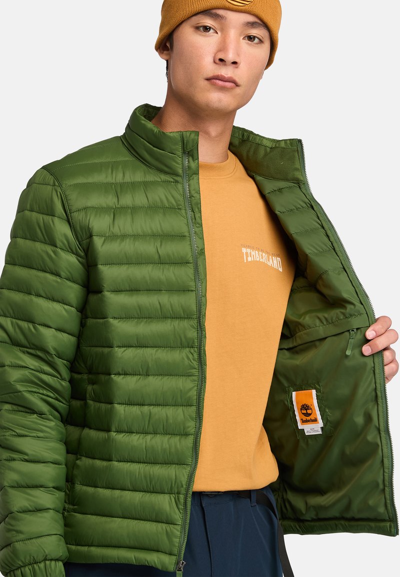 Zielona kurtka puchowa z poziomym pikowaniem, pełnym zapięciem na zamek i miękką podszewką. Wewnętrzna kieszeń z metką z logo Timberland.