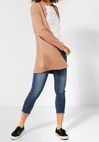 Vaaleanruskea neule-cardigan, jossa on avoin etuosa, pitkät hihat ja pehmeä tekstuuri, yhdistetty valkoiseen graafiseen t-paitaan ja sinisiin farkkuihin. Mustat tennarit täydentävät asun.