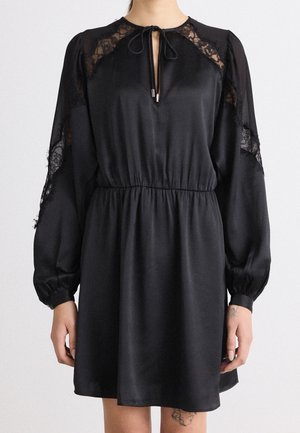 Robe noire à manches longues avec panneaux en dentelle sur les épaules et les manches, taille élastique, décolleté en trou de serrure noué, portée par une personne avec des tatouages sur les mains et les jambes.