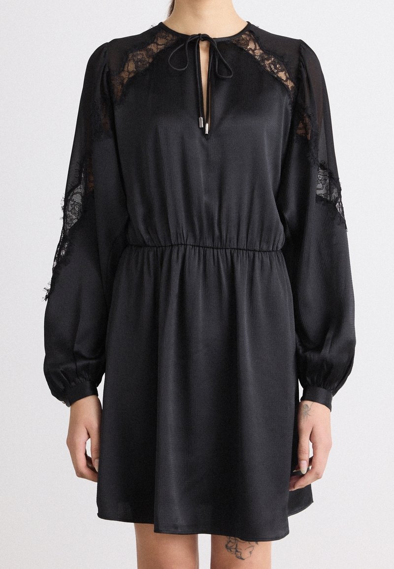 Robe noire à manches longues avec panneaux en dentelle sur les épaules et les manches, taille élastique, décolleté en trou de serrure noué, portée par une personne avec des tatouages sur les mains et les jambes.