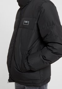 Veste matelassée noire avec col montant, poches latérales et un écusson logo rectangulaire. Texture lisse avec des sections matelassées.