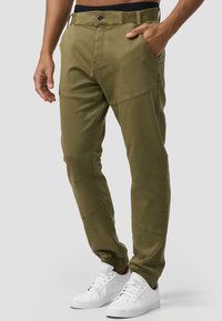 Pantalones ajustados color verde oliva con detalles de patchwork, con cierre de botón, bolsillos laterales y una cinturilla cómoda, combinados con zapatillas blancas.