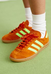 Orange mockaskinnsneakers med neongröna ränder, rund tå och gummisula. Har röda snören och vadderad vrist.