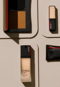 Bottiglia di fondotinta in vetro opaco con tappo nero e rosso, compatto con polvere marrone, una piccola custodia per rossetto e un tubo nero con dettagli rossi.