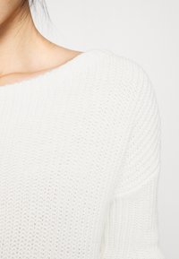 Witte gebreide trui met een brede halslijn, ribbelpatroon en een ontspannen pasvorm. De stof lijkt zacht en licht van gewicht.