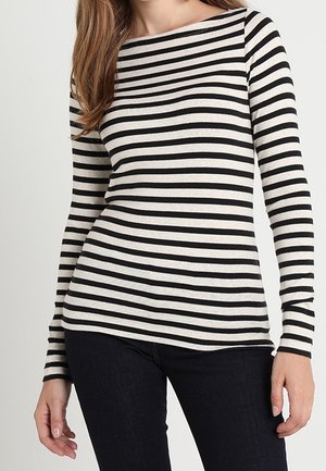 Woman wearing a long-sleeve black and white horizontal striped top and dark pants, standing against a plain light background.