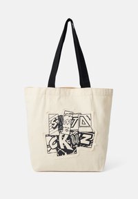 Sac tote en toile crème avec des poignées noires. Le devant présente un design graphique avec du texte et des motifs superposés, ajoutant texture et détail.