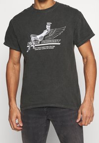 Černé grafické tričko vyrobené z bavlny má bíle potisknutý design okřídlené postavy s textem, kulatý výstřih a krátké rukávy.