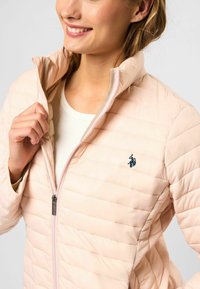 Vadderad ljusrosa jacka med hög krage, med horisontell quiltning och en liten marinblå logotyp på bröstet, öppnad för att avslöja innerskiktet.
