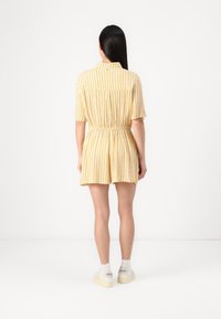 Rompers court jaune à rayures avec des manches courtes et une taille élastique, doté d'un col et d'une coupe décontractée. Porté avec des baskets blanches.