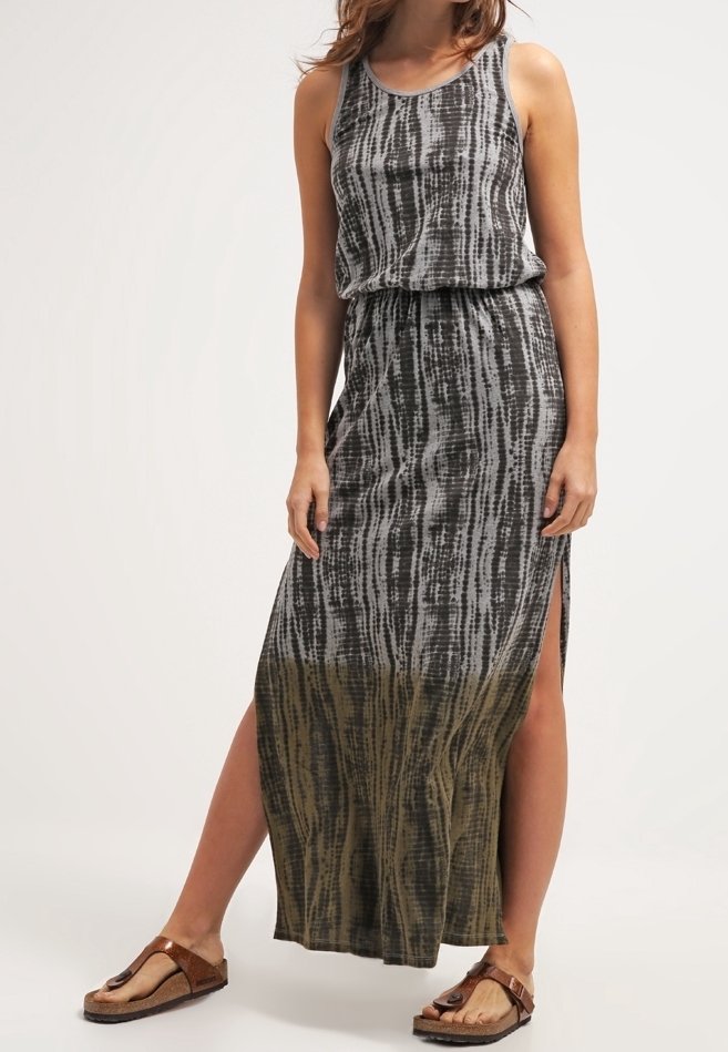 Femme portant une robe maxi sans manches tie-dye noire et grise avec des fentes latérales, associée à des sandales marron à boucle, debout devant un fond uni.