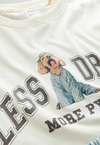Šviesiai kreminis megztinis su šunelio grafika, apsirengusio mėlynai dryžuotais pižamais. Užrašas sako "MAŽIAU DRAMOS, DAUGIAU PIŽAMŲ" dideliu, juodu šriftu.