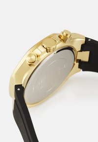 Reloj cronógrafo de tono dorado con tres botones de pulsación, correa de silicona negra y una elegante caja redonda. Presenta una superficie lisa con respaldo plateado pulido.