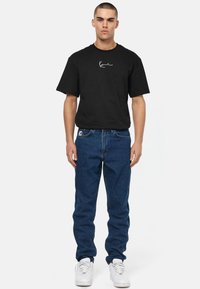 Μαύρο κοντομάνικο μπλουζάκι με λευκό λογότυπο, συνδυασμένο με μπλε τζιν. Μοντέλο που φορά λευκά αθλητικά παπούτσια, στέκεται μπροστά σε ουδέτερο φόντο.